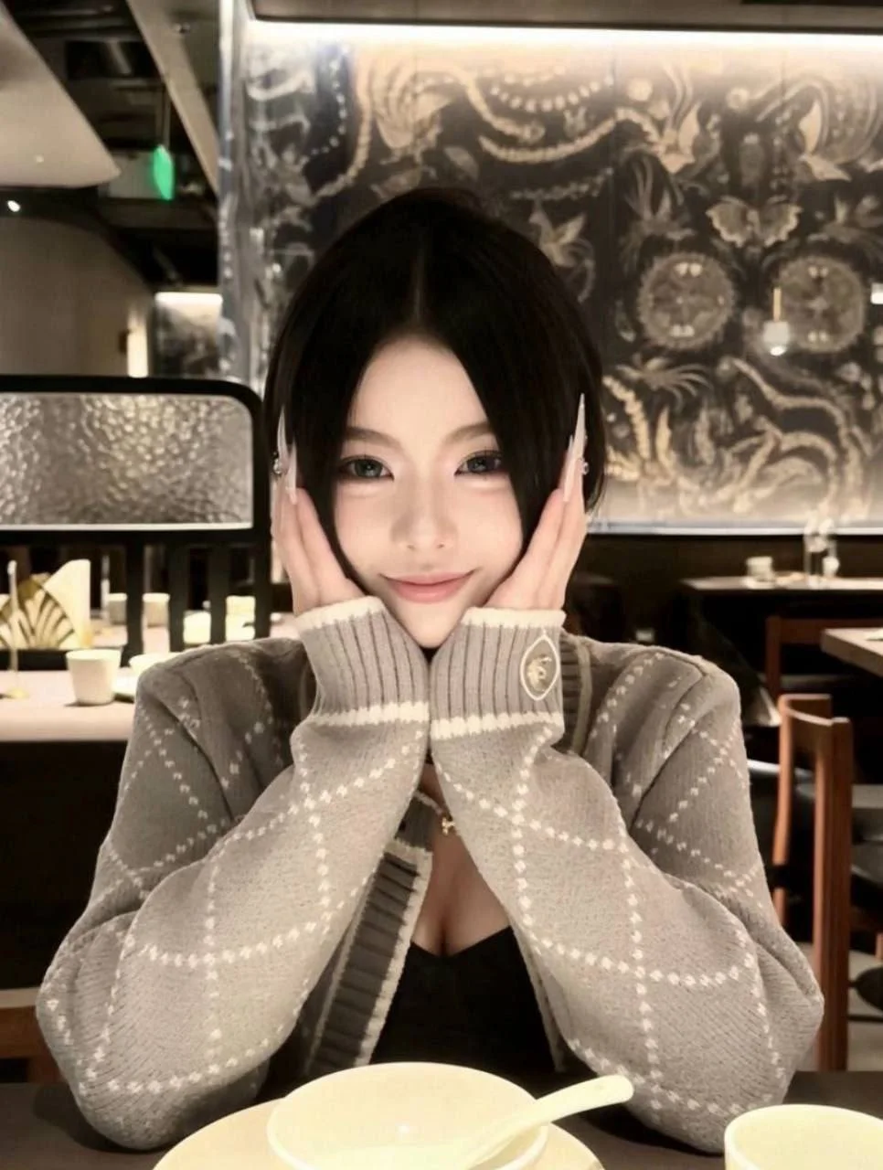 餐厅里黑发女孩双手托脸微笑，身后是花纹墙